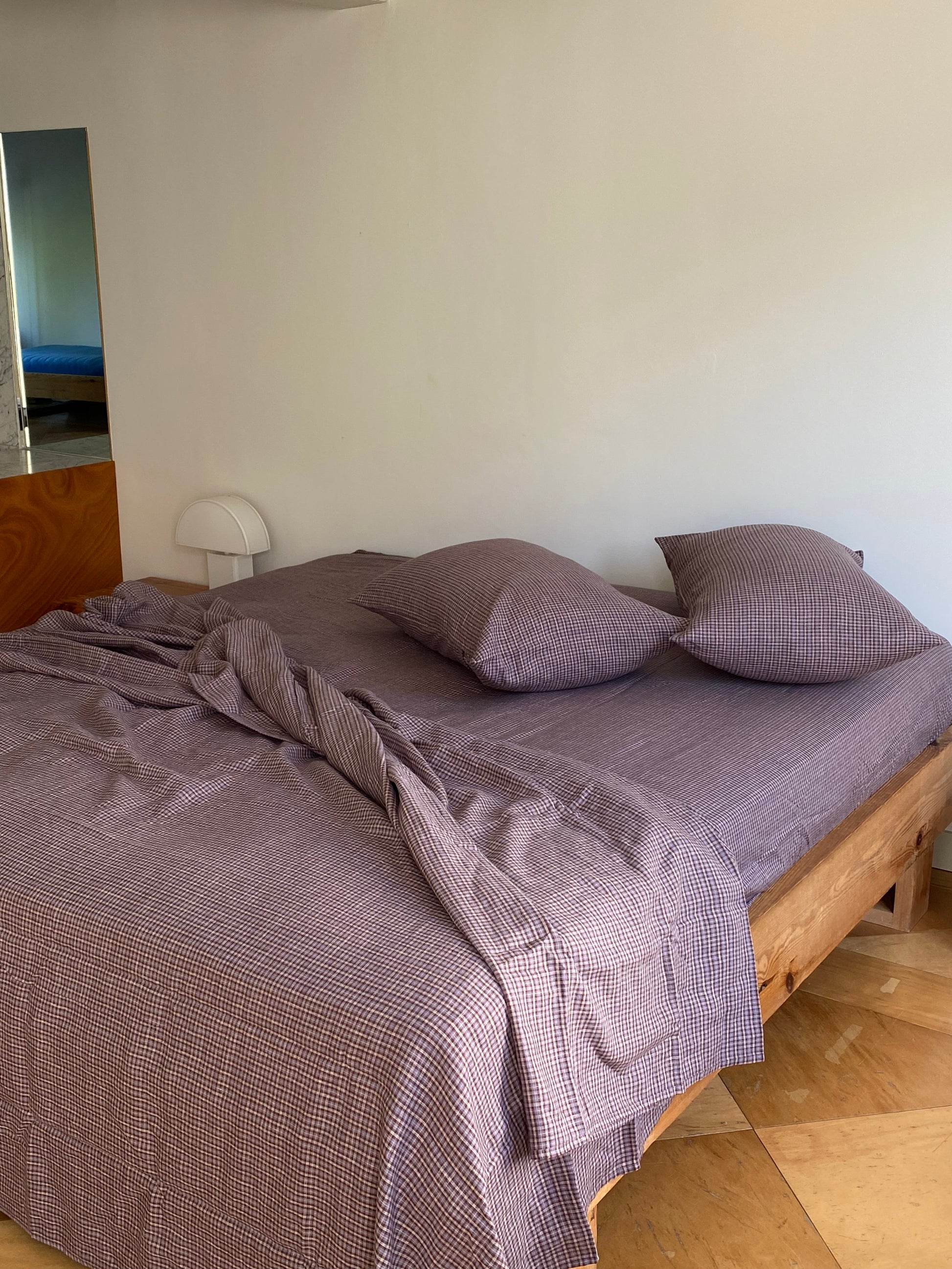 A neatly made wooden bed is dressed in purple checkered sheets and two Pillow slip set - Russet Check. The sheet is slightly rumpled, with a small white lamp on the bedside table against a plain wall.