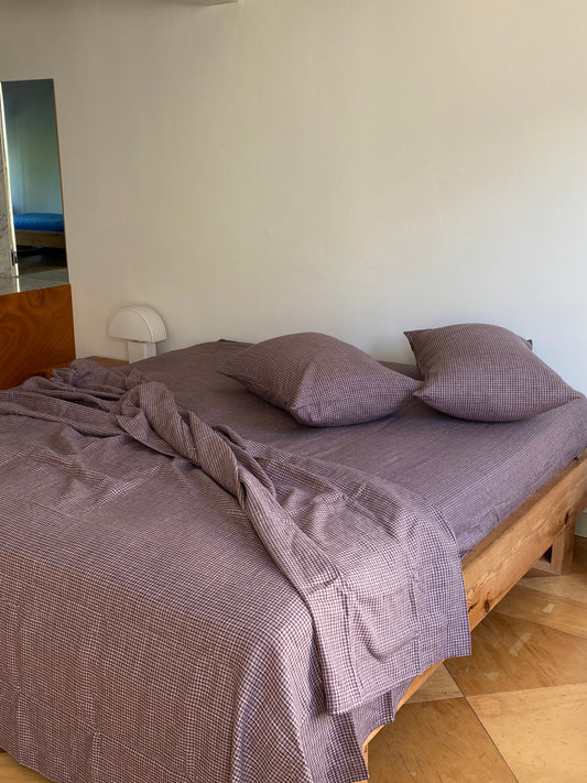 A neatly made bed with a wooden frame, purple checkered sheets, and two Pillow slip set - Russet Check pillowcases in a bright room with light-colored walls and a mirror reflecting another bed.