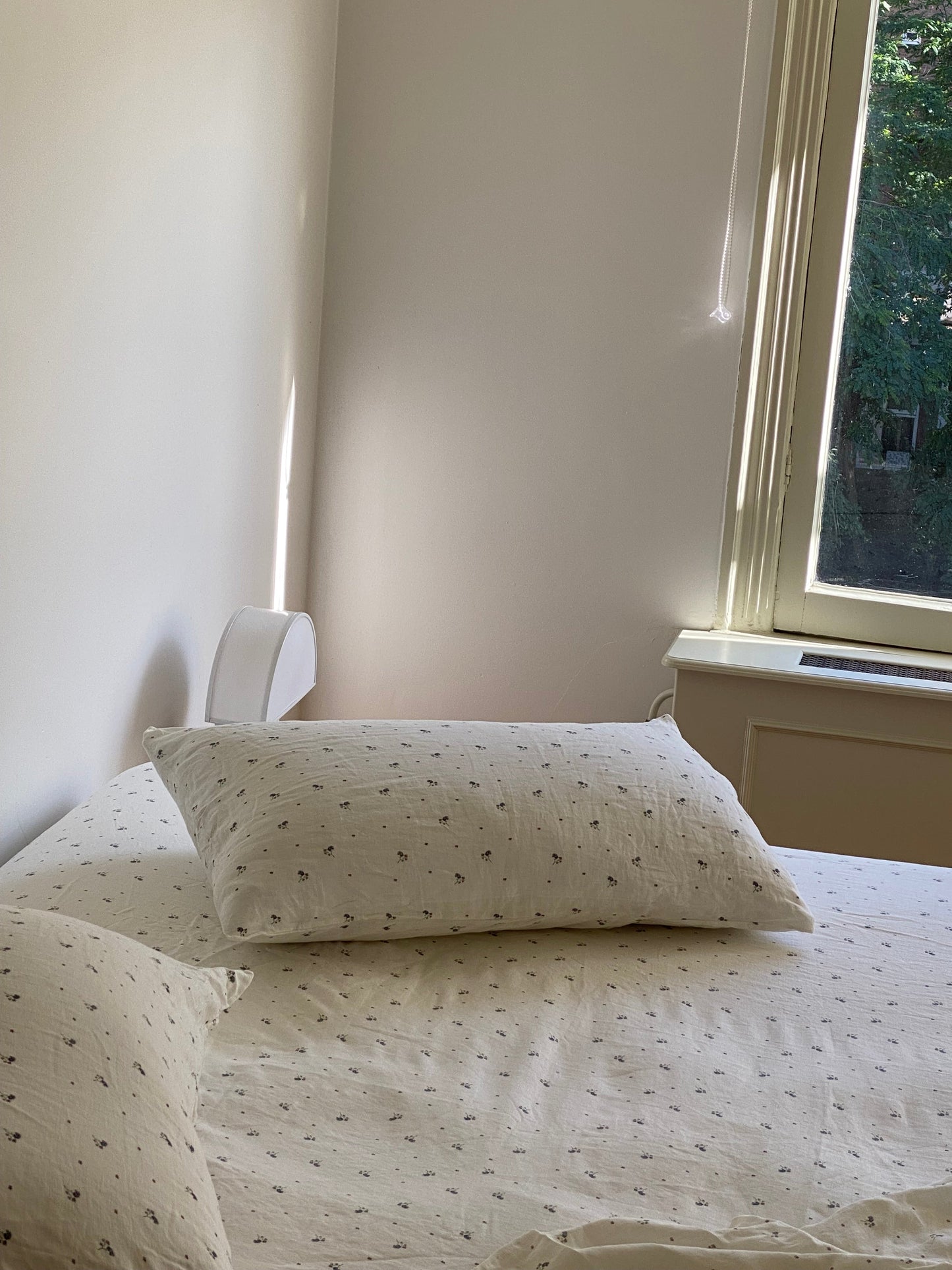 A neatly made bed with white sheets and Pillow Slips featuring small dark patterns sits next to a sunlit window with trees outside, as soft light fills the serene, minimal room.