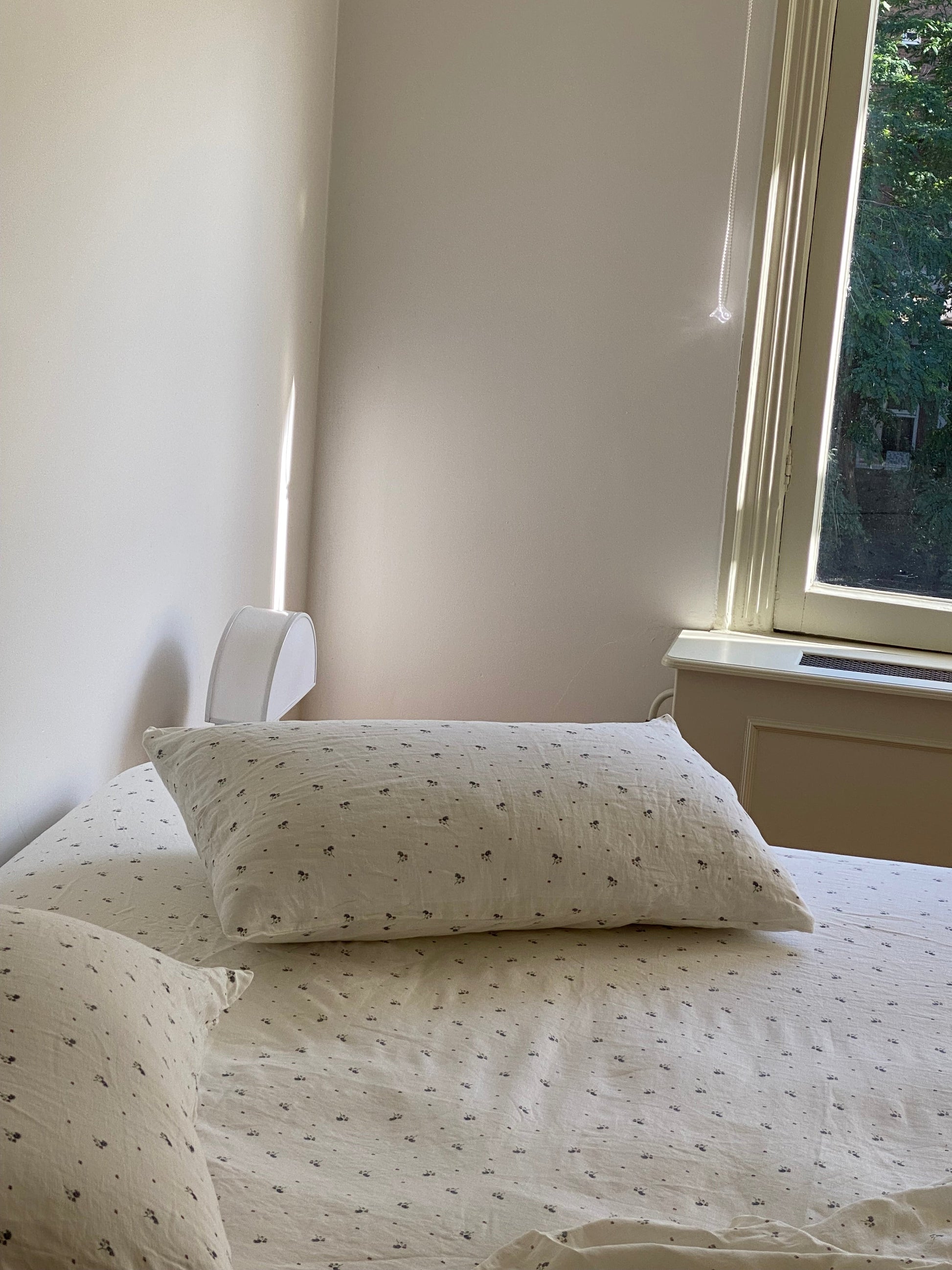 A neatly made bed with white sheets and Pillow Slips featuring small dark patterns sits next to a sunlit window with trees outside, as soft light fills the serene, minimal room.