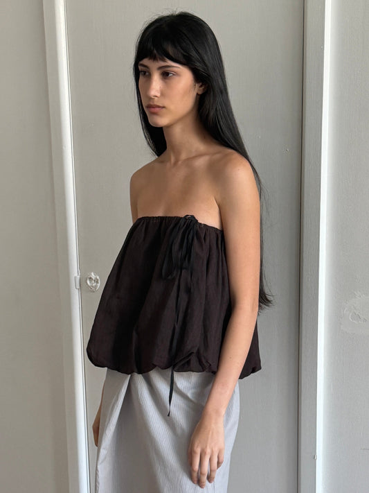 A woman with dark hair and a fringe stands in profile in a room wearing the Bubble Top - Brown Black Stripe by Deiji Studios, a strapless dark brown bubble-hem top with a drawcord tie, over a pale grey skirt.