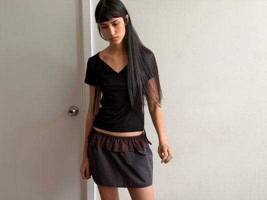 A woman with dark hair and a fringe stands looking down, wearing the Tie Back Tee - Black by Deiji Studios, a fitted black tee with a sweetheart neckline and back ties, with a brown flounce mini skirt.