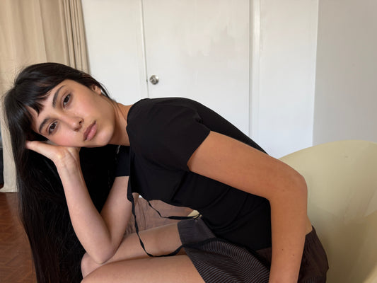 A woman with dark hair and a fringe reclines in a chair wearing the Tie Back Tee - Black by Deiji Studios, a fitted black tee with a sweetheart neckline and back ties, with a striped mini skirt.