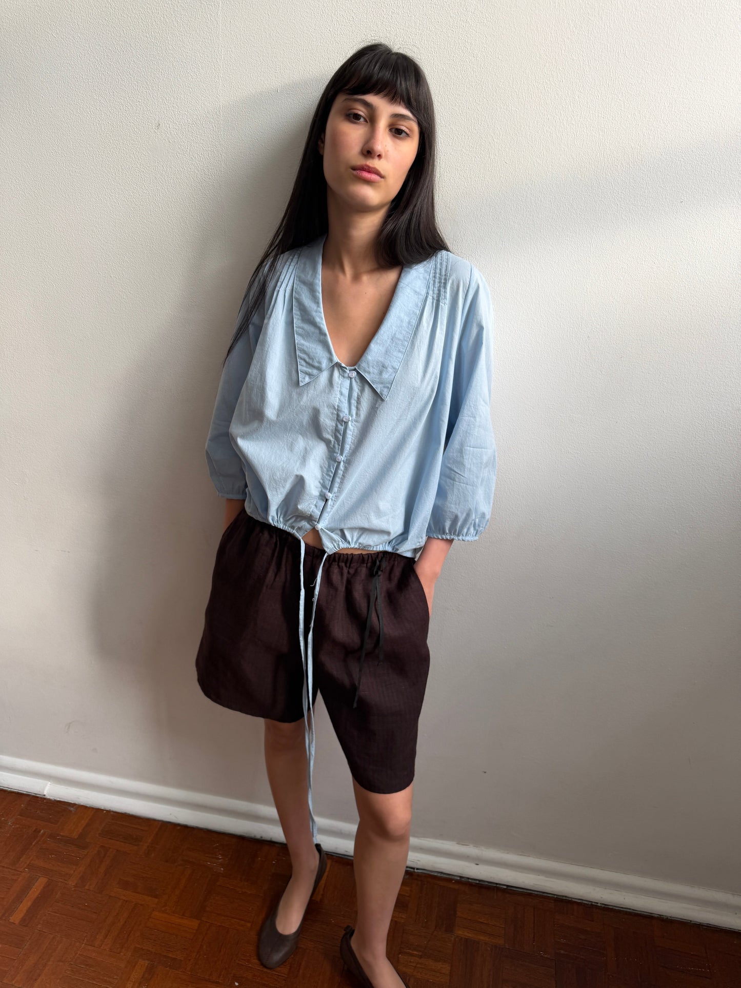 A woman with dark hair and a fringe stands against a wall wearing the Shoulder Tucks Top - Baby Blue by Deiji Studios, a baby blue blouse with a large pointed collar, buttoned V-placket and gathered hem, with brown shorts and flats.