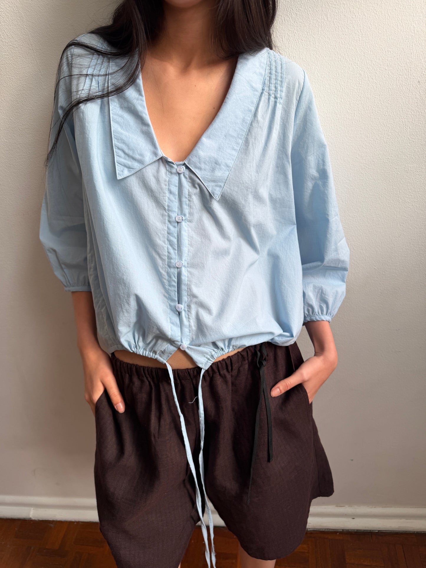 A cropped view of the Shoulder Tucks Top - Baby Blue by Deiji Studios, a baby blue blouse with a large pointed collar, buttoned V-placket and drawcord hem, worn with brown shorts against a pale wall.