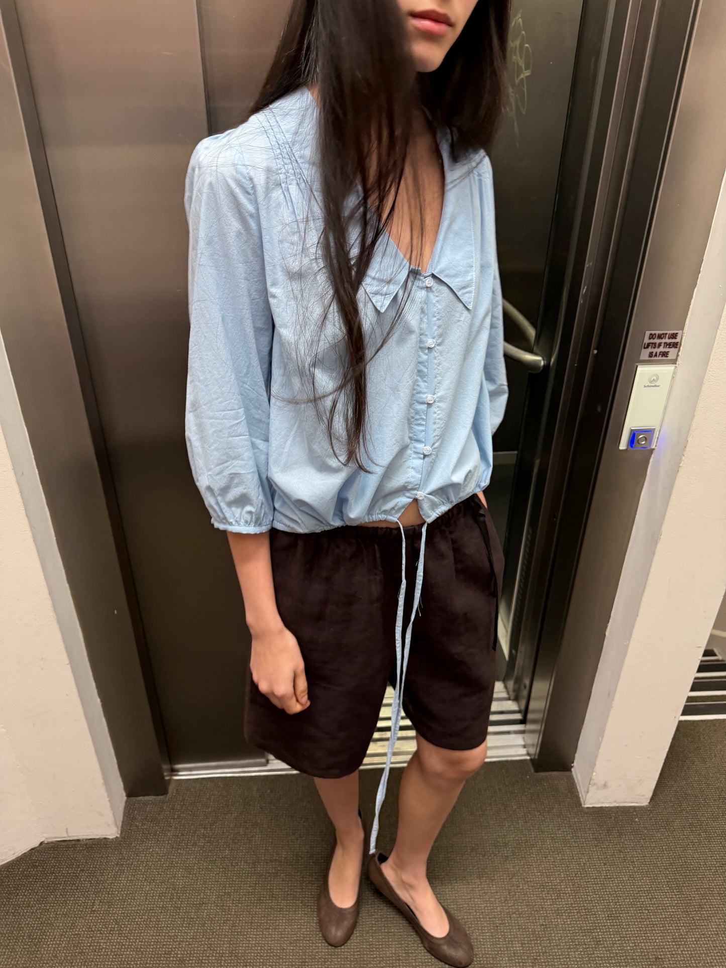 A woman with long dark hair stands in front of a lift wearing the Shoulder Tucks Top - Baby Blue by Deiji Studios, a baby blue blouse with a large pointed collar and gathered hem, with brown shorts and flats.