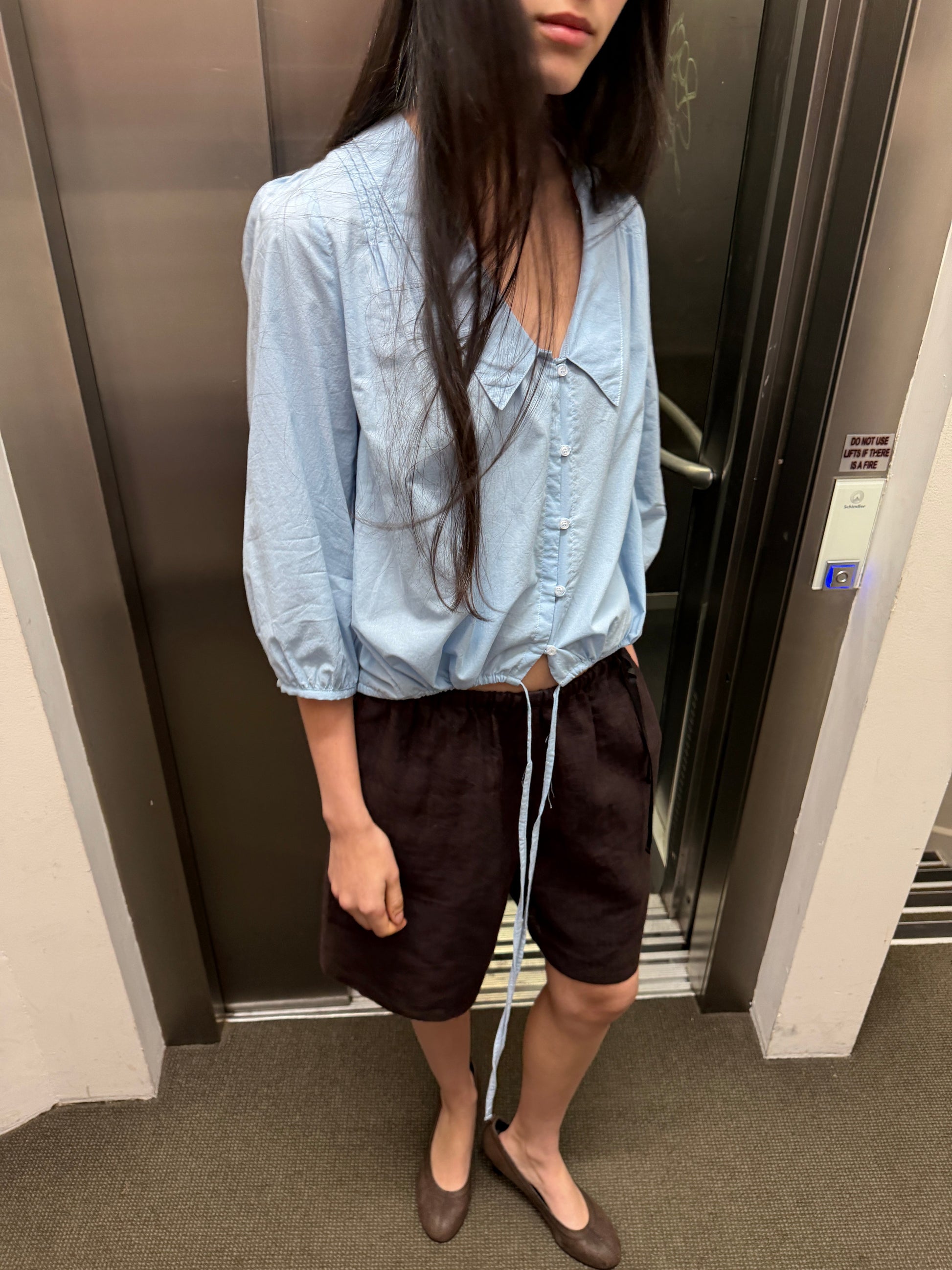 A woman with long dark hair stands in front of a lift wearing the Shoulder Tucks Top - Baby Blue by Deiji Studios, a baby blue blouse with a large pointed collar and gathered hem, with brown shorts and flats.