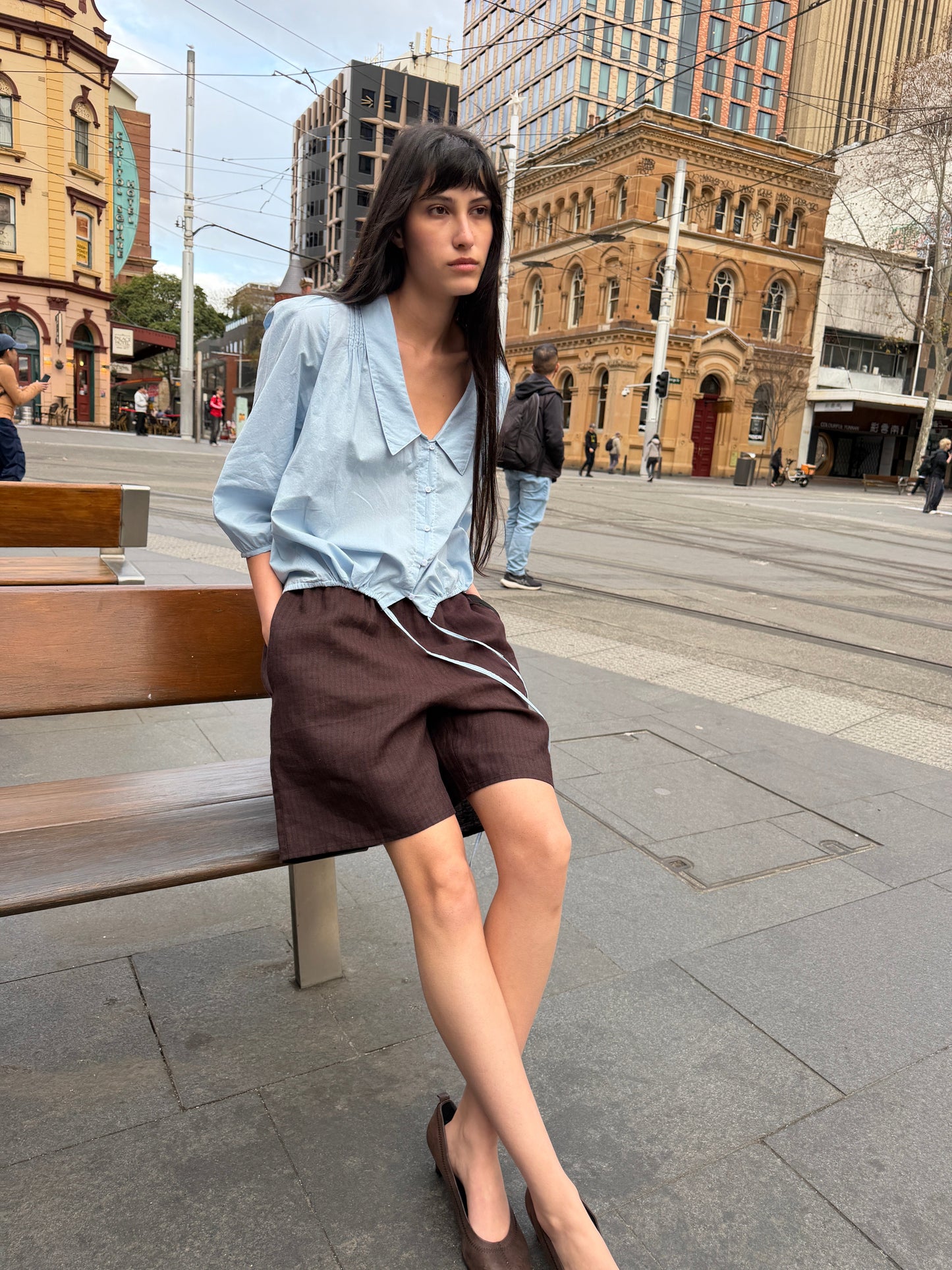 A woman with long dark hair sits on a bench in a city square wearing the Shoulder Tucks Top - Baby Blue by Deiji Studios, a baby blue blouse with a large pointed collar, and brown shorts.