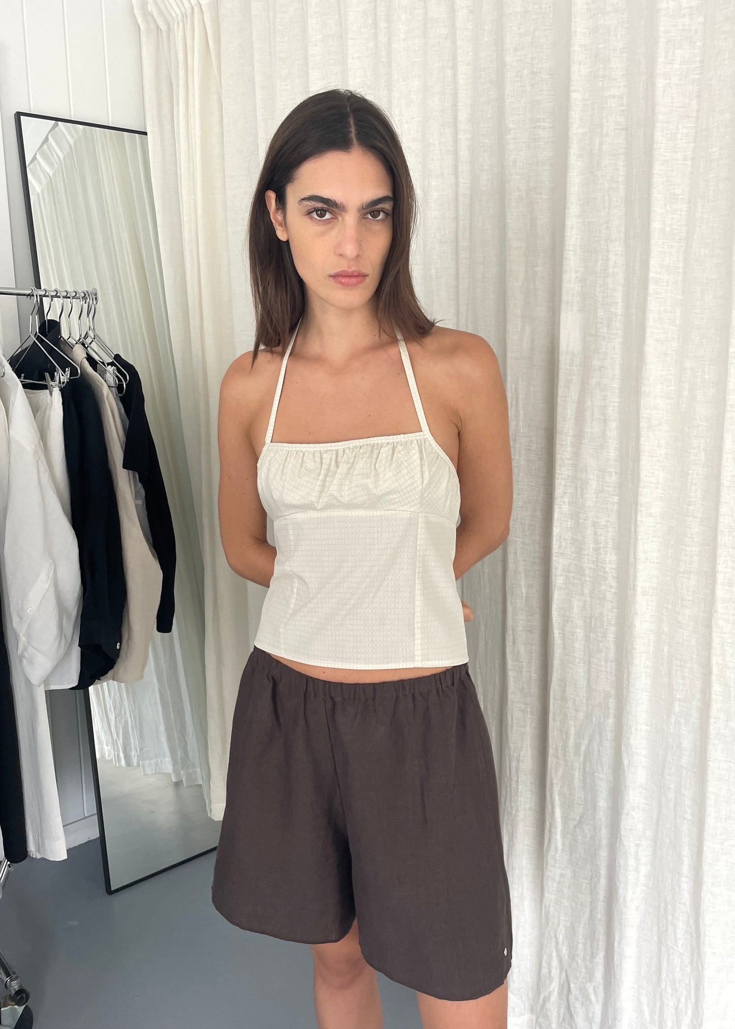 A woman with straight brown hair stands indoors against white curtains, wearing a white Strap Top and loose brown shorts, hands behind her back. Clothing racks and a mirror are in the background.