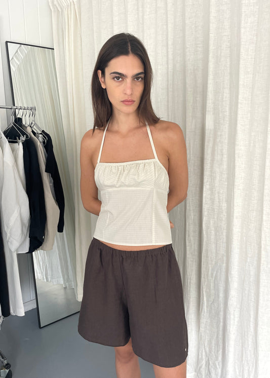 A woman with straight brown hair stands indoors against white curtains, wearing a white Strap Top and loose brown shorts, hands behind her back. Clothing racks and a mirror are in the background.