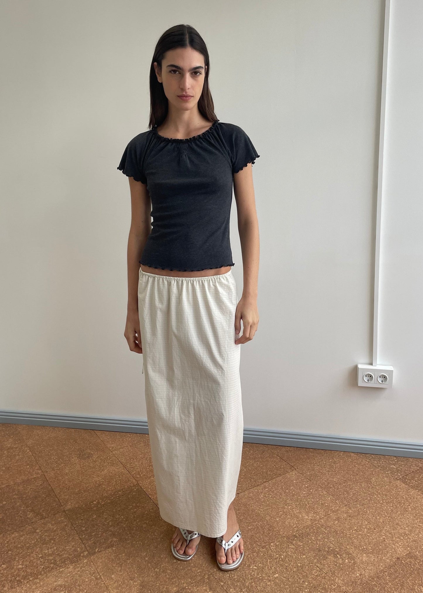 A woman with long dark hair and a neutral expression stands indoors against a plain wall, wearing a black short-sleeve top, flip-flops, and the Mid Day Skirt - Bone Check. An electrical outlet is visible on the wall behind her.