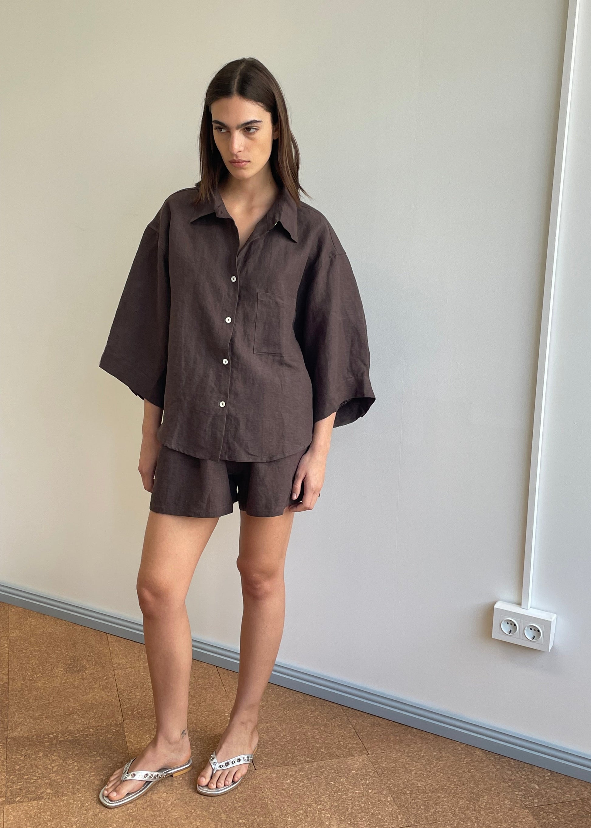 A woman with straight brown hair and a neutral expression stands indoors against a plain wall, wearing the 03 Set - Bark: a loose-fitting organic linen shirt and matching boxer-style shorts, paired with sandals.
