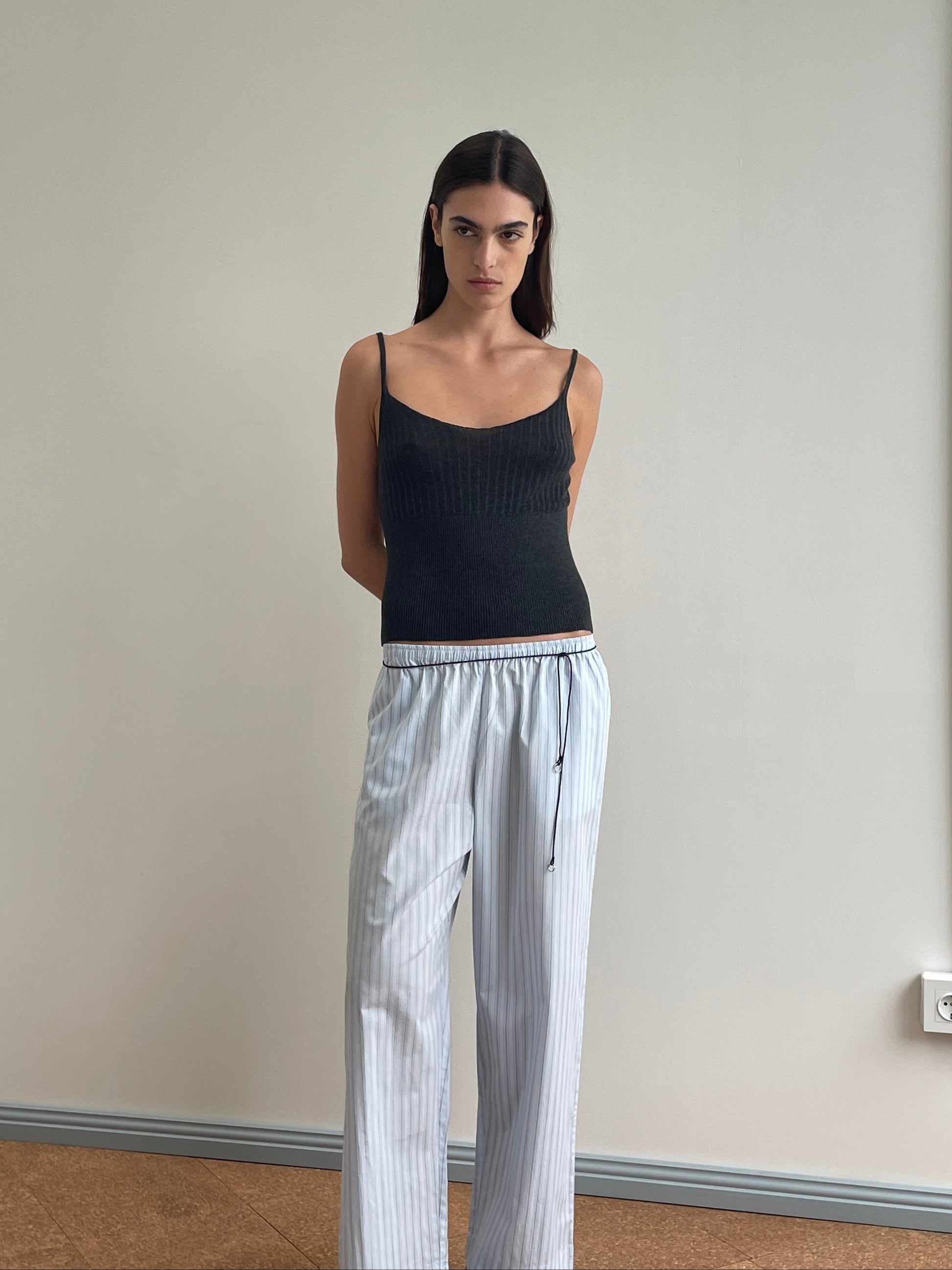A woman with straight dark hair stands indoors against a plain wall, wearing a black sleeveless top and the Ease Trouser - Mineral Stripe by Deiji Studios in organic cotton, her hands resting behind her back.