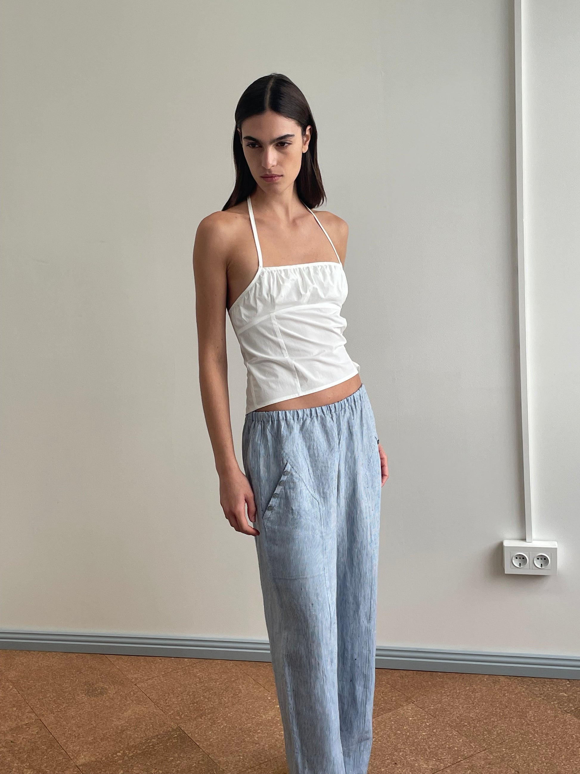 A woman stands indoors against a plain wall, wearing the Deiji Studios Strap Top - White and light blue loose organic cotton pants, her hands in her pockets and a neutral expression.