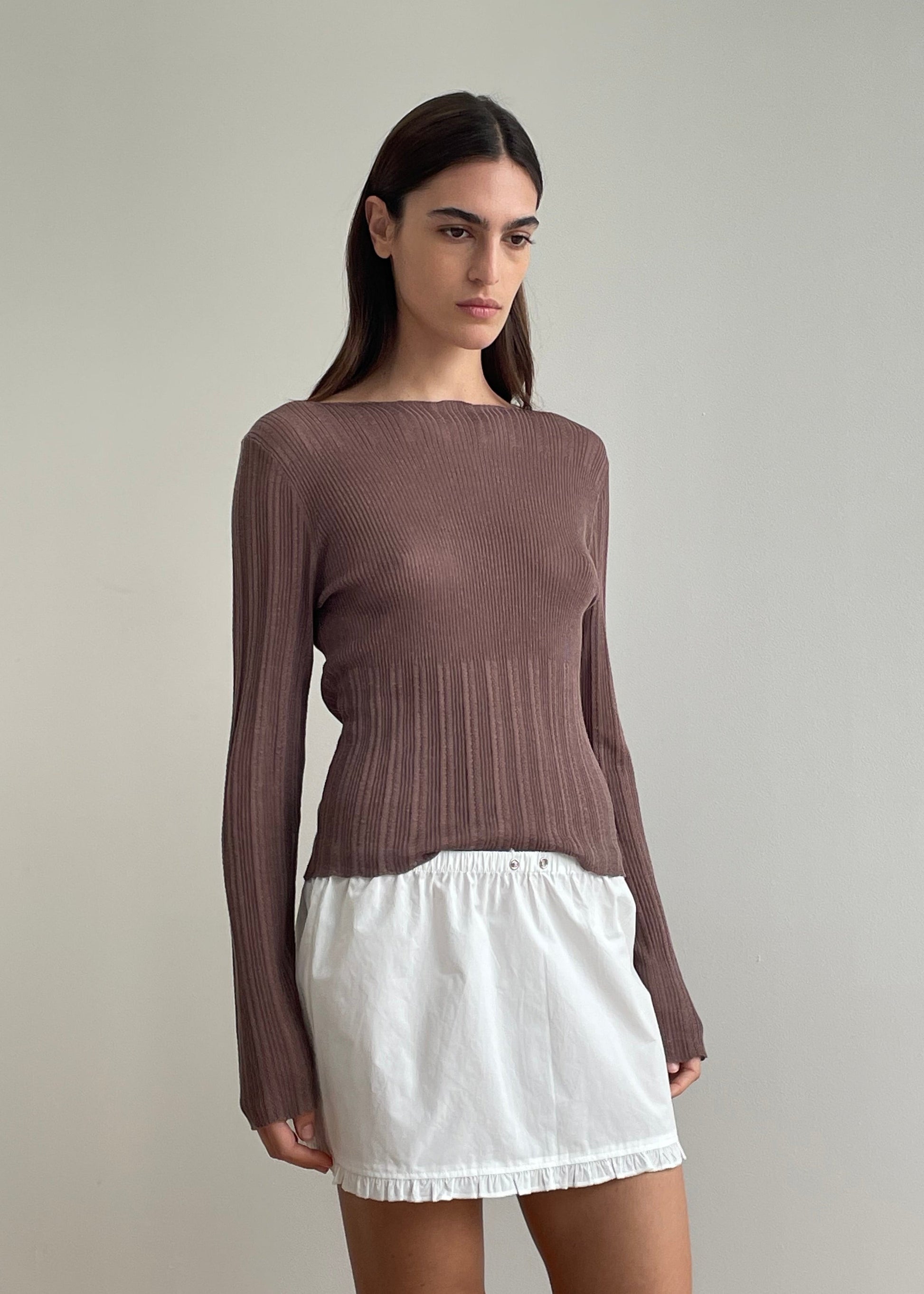 A woman with straight dark hair and a neutral expression stands against a light wall, wearing the Open Work Longsleeve - Hazel paired with a short white skirt.