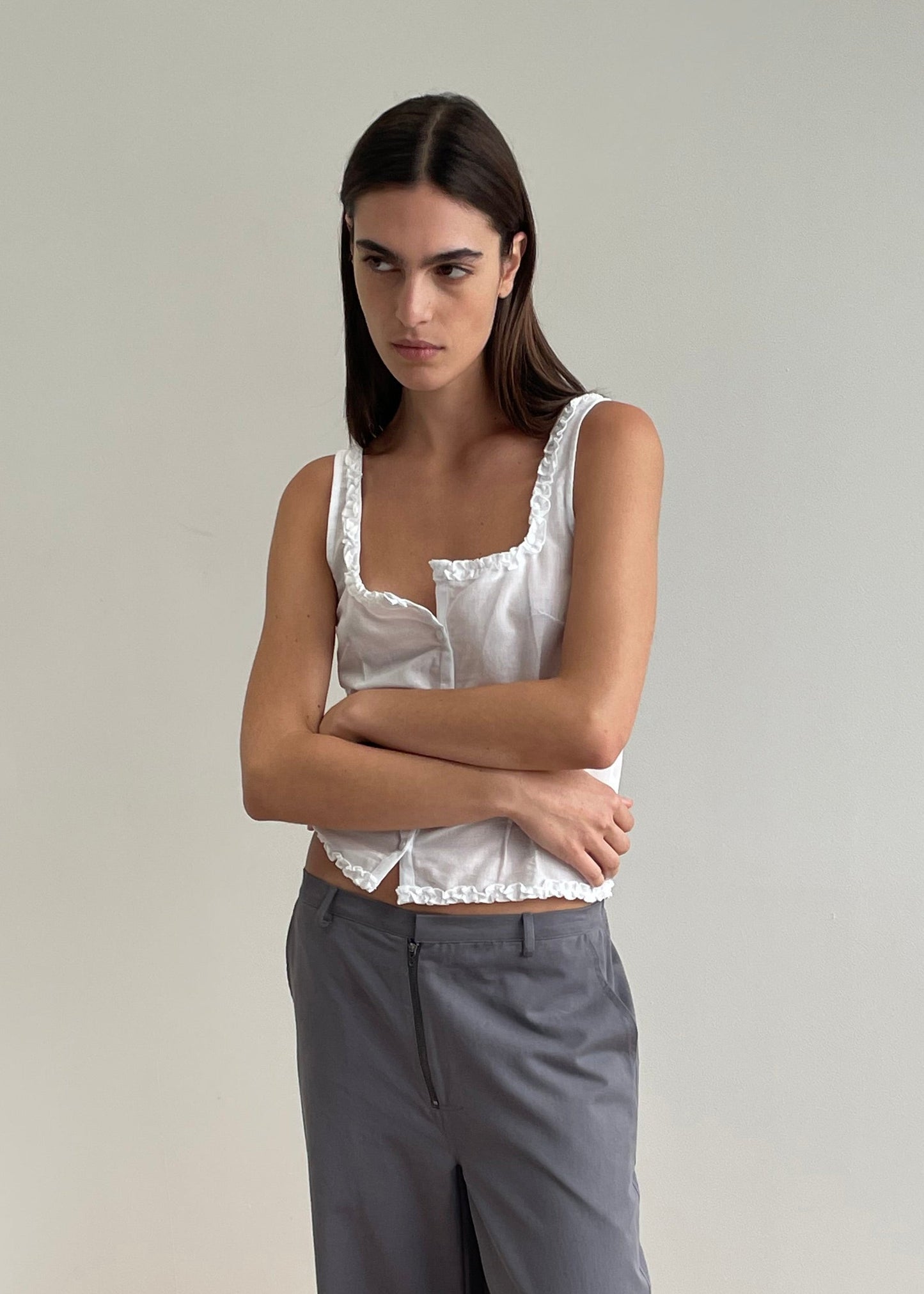 A woman with long dark hair stands against a plain light wall, wearing a white Button Frill Top from Deiji Studios and gray pants, her arms crossed and a neutral expression on her face.
