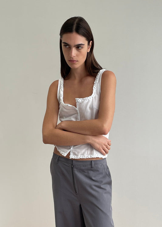 A woman with long dark hair stands against a plain light wall, wearing a white Button Frill Top from Deiji Studios and gray pants, her arms crossed and a neutral expression on her face.