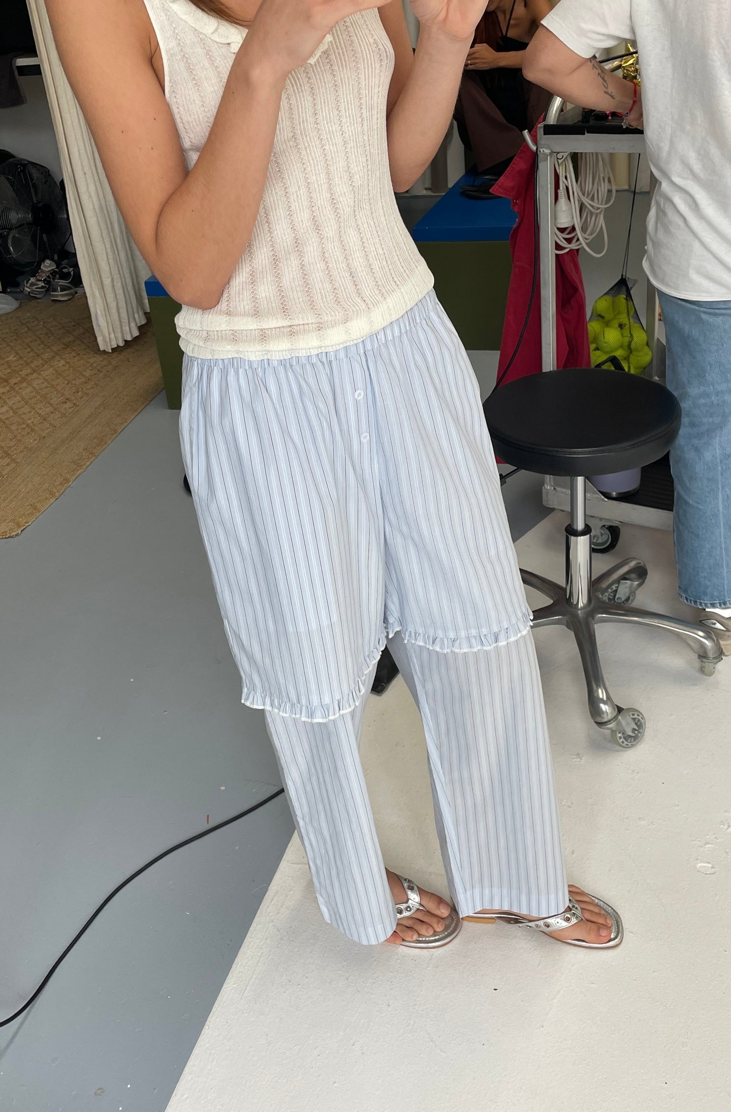 A person wears the Flounce Short - Mineral Stripe sleeveless knitted top with loose striped pants and sandals, standing indoors on a light floor. Only their upper body and legs are visible, with people and office items in the background.