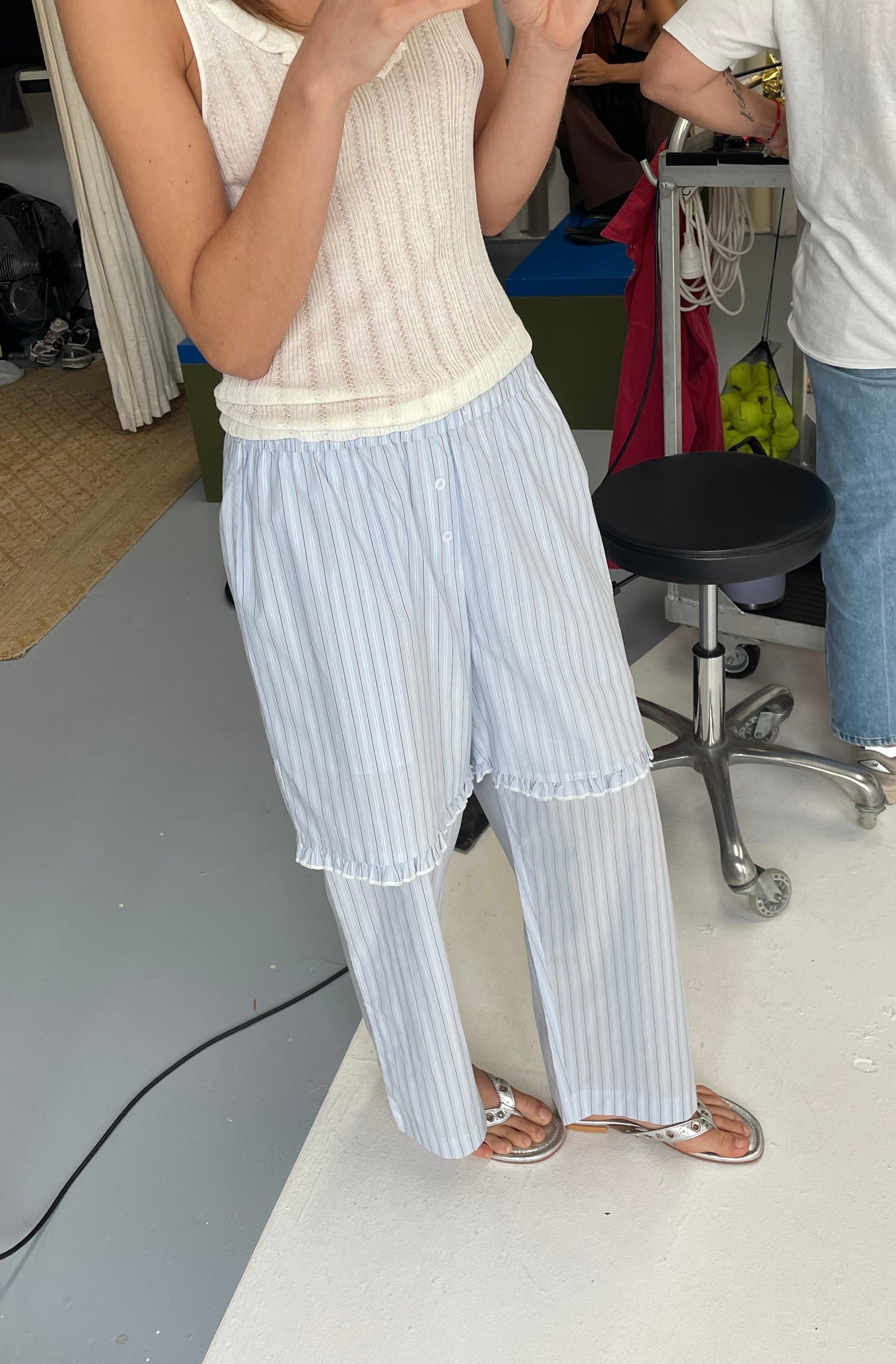 A person wears the Flounce Short - Mineral Stripe sleeveless knitted top with loose striped pants and sandals, standing indoors on a light floor. Only their upper body and legs are visible, with people and office items in the background.
