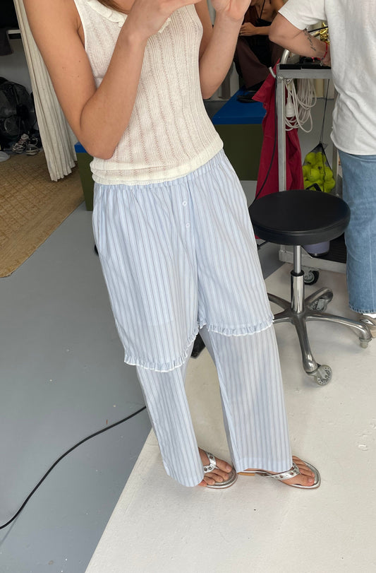 A person wears the Flounce Short - Mineral Stripe sleeveless knitted top with loose striped pants and sandals, standing indoors on a light floor. Only their upper body and legs are visible, with people and office items in the background.