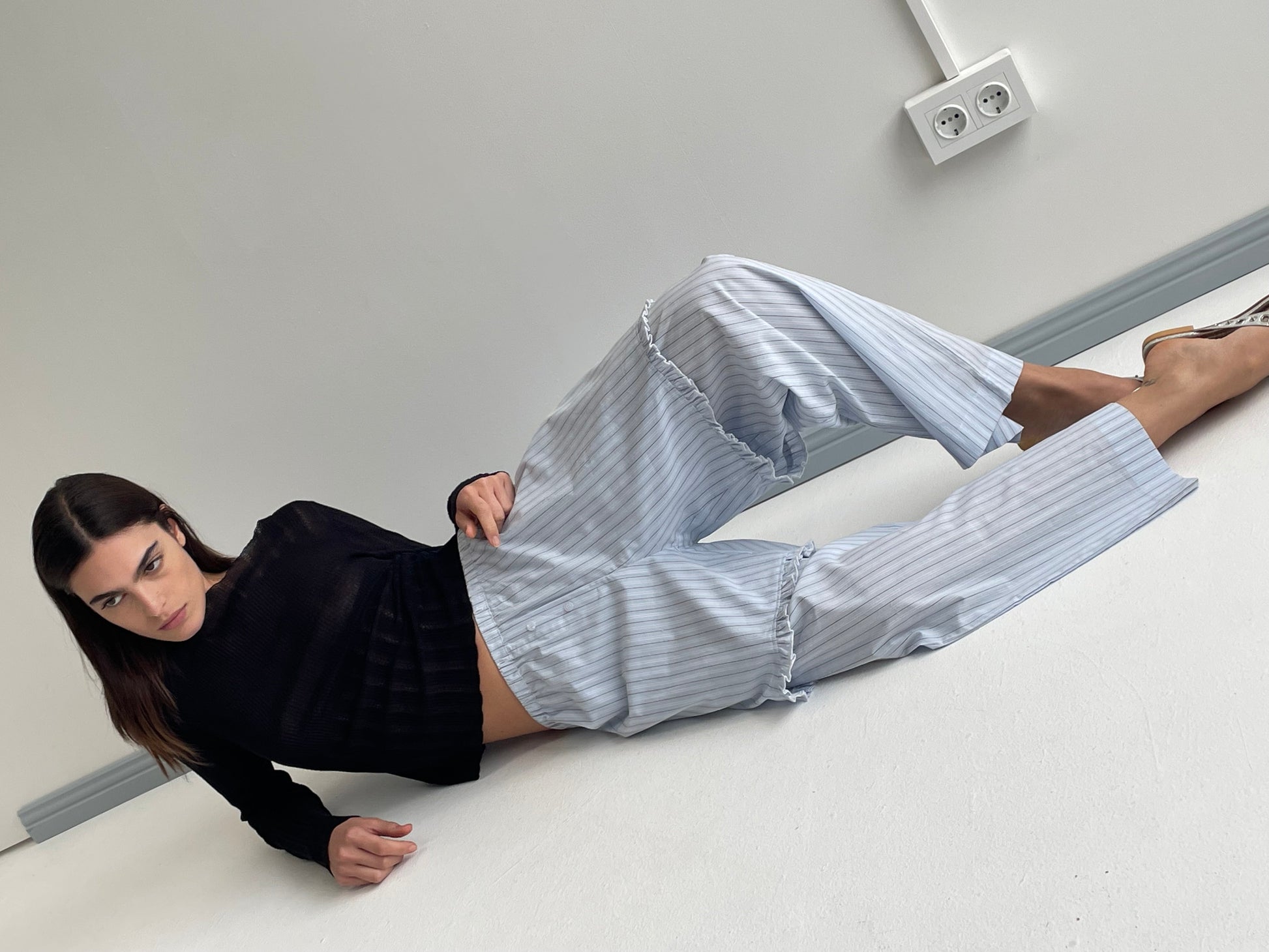 A woman with straight dark hair lies on the floor by a white wall, wearing a black long-sleeve top and the Flounce Short – Mineral Stripe. One leg is bent as she looks at the camera, near a wall outlet.
