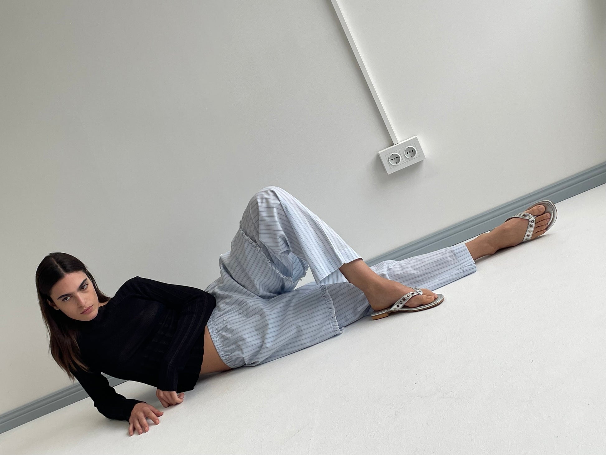 A woman in a black long-sleeve top and Ease Trouser - Mineral Stripe by Deiji Studios, made from organic cotton, reclines on the floor against a white wall with one knee bent and the other leg extended, gazing at the camera.