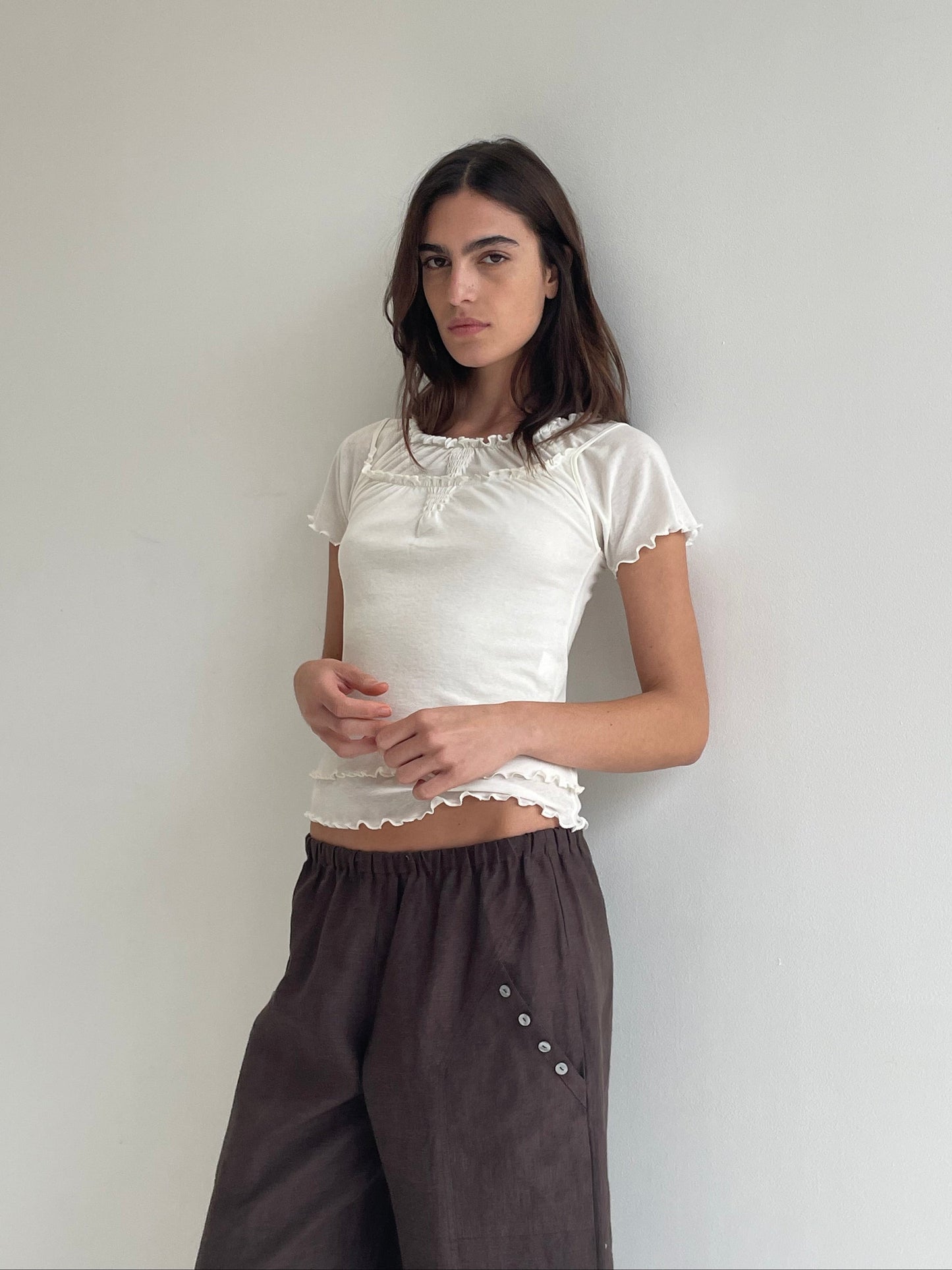 A woman stands against a plain light wall, wearing the Flounce Tee - Cream by Deiji Studios with ruffled edges and loose brown pants with button details. She looks at the camera with a neutral expression.