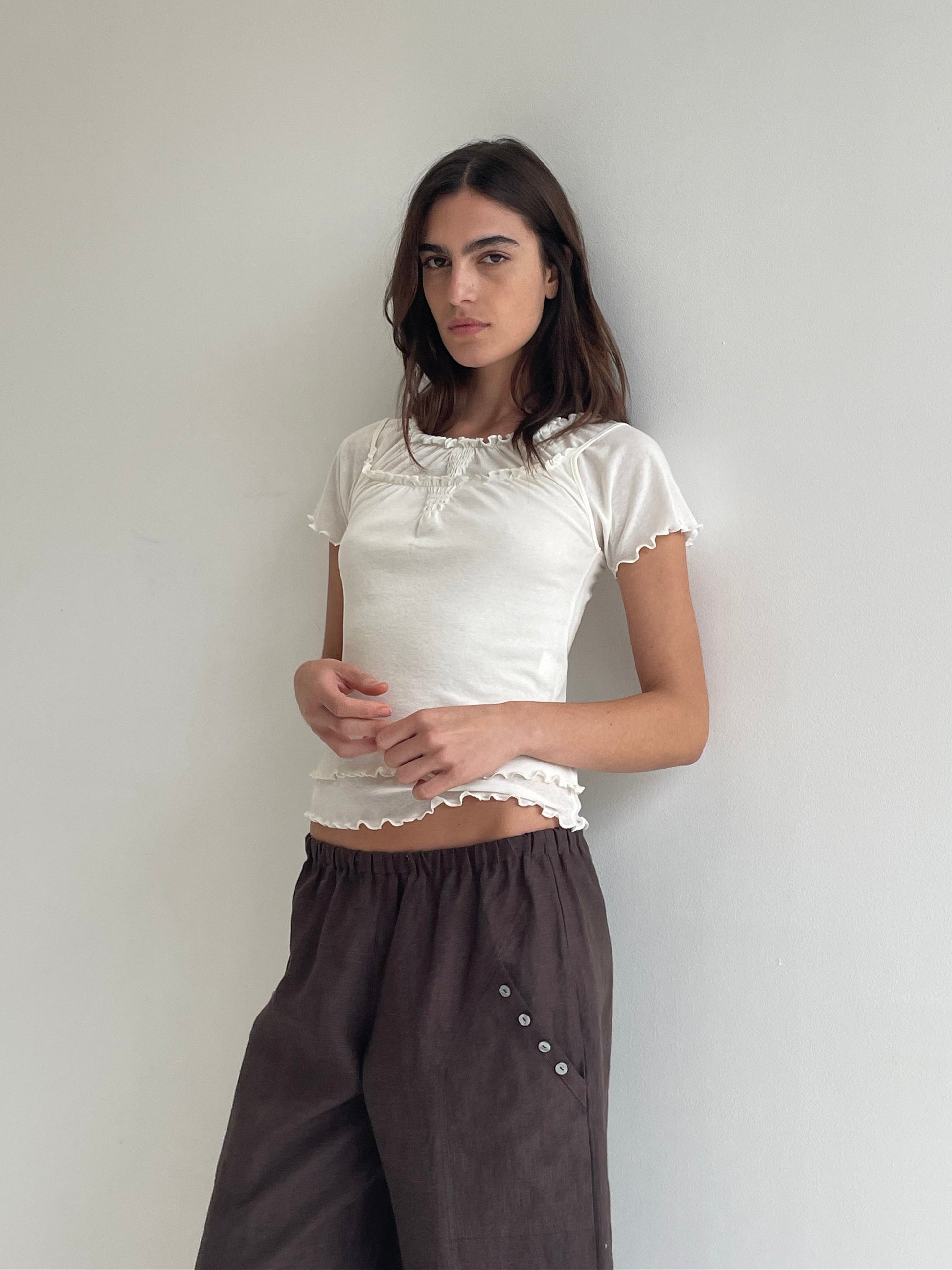 A woman stands against a plain light wall, wearing the Flounce Tee - Cream by Deiji Studios with ruffled edges and loose brown pants with button details. She looks at the camera with a neutral expression.