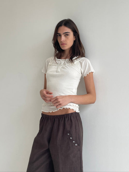 A woman stands against a plain light wall, wearing the Flounce Tee - Cream by Deiji Studios with ruffled edges and loose brown pants with button details. She looks at the camera with a neutral expression.