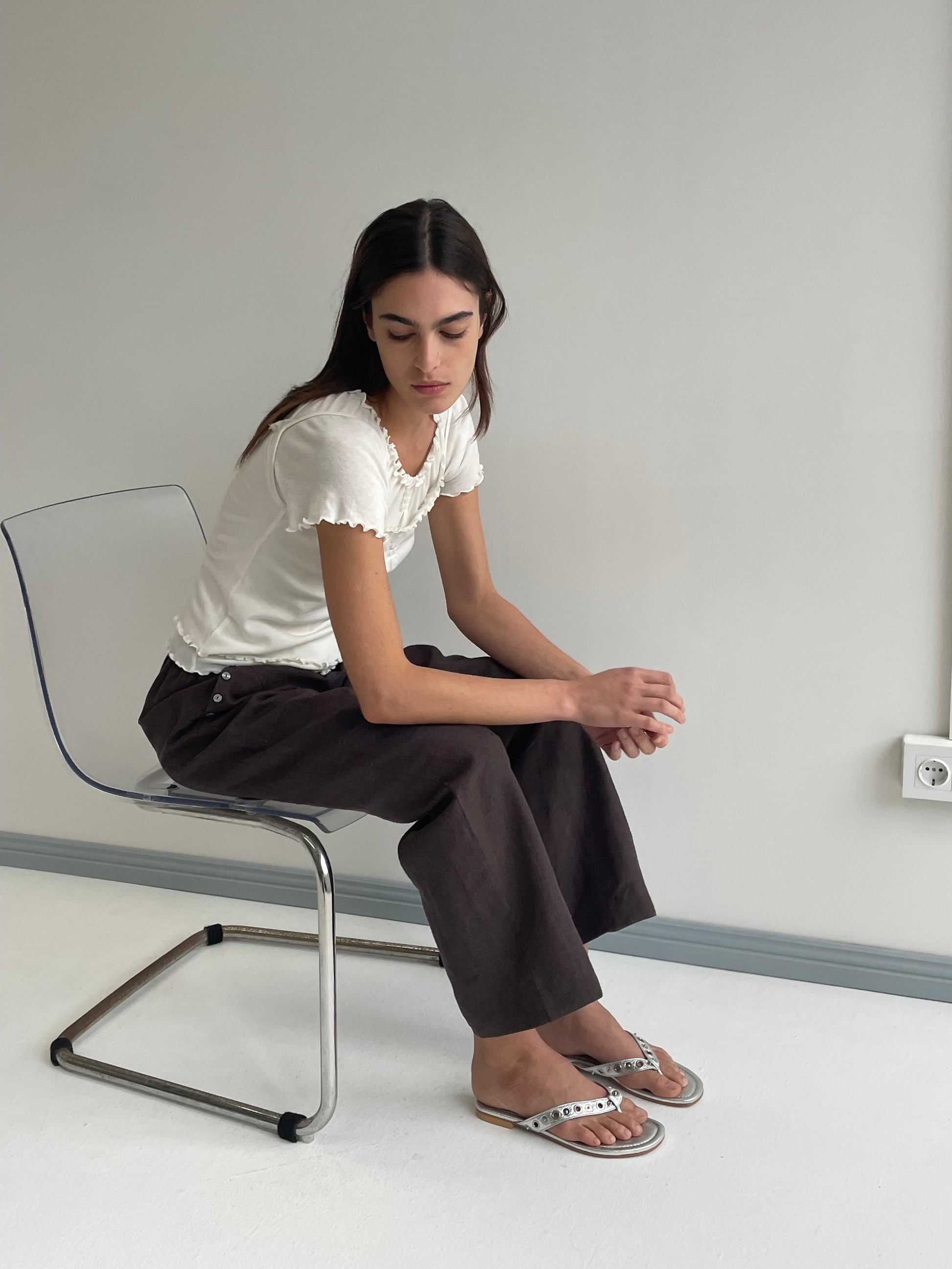 A woman with long dark hair sits on a transparent chair, wearing a white short-sleeve top and the Angle Pocket Pant in Bark—organic linen pants with a relaxed mid-waist fit—paired with white sandals, gazing down against a plain light wall.
