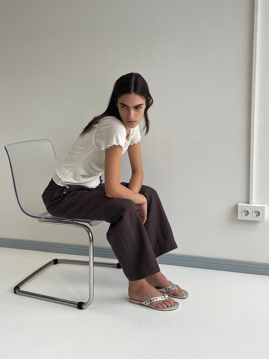 A woman with long dark hair sits sideways on a transparent chair, wearing the Flounce Tee - Cream by Deiji Studios, loose dark brown pants, and white sandals in a minimalist room with light walls and a power outlet.