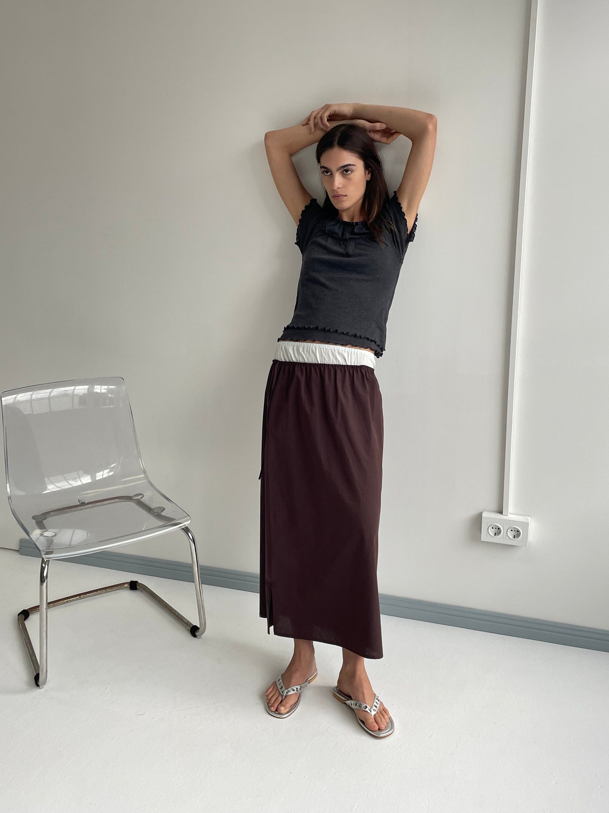 A model poses with arms raised wearing a dark grey frilled crop top, the Mid Day Skirt - Umber featuring an A-line silhouette and burgundy elasticated waistband, and silver sandals beside a clear acrylic chair in a minimalist white room.