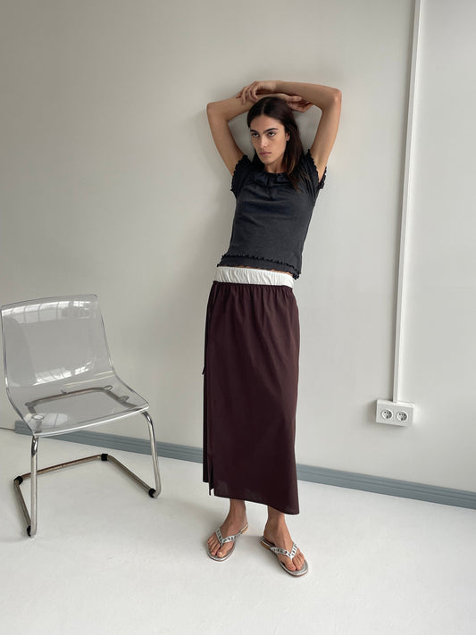 A model poses with arms raised wearing a dark grey frilled crop top, the Mid Day Skirt - Umber featuring an A-line silhouette and burgundy elasticated waistband, and silver sandals beside a clear acrylic chair in a minimalist white room.