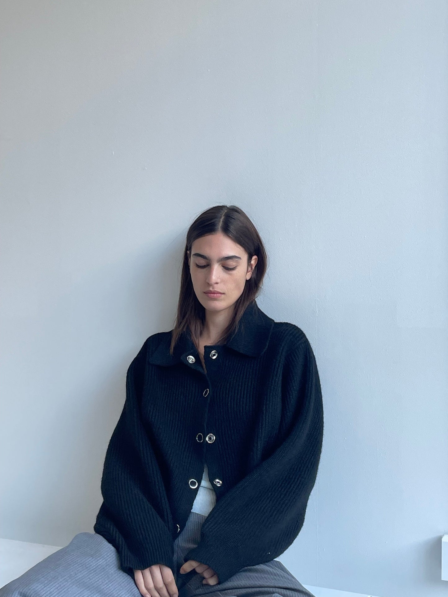 A woman with long brown hair sits against a light gray wall, wearing the Stud Cardi Knit - Black in an oversized fit and gray pants. She looks down, her expression calm and serene.