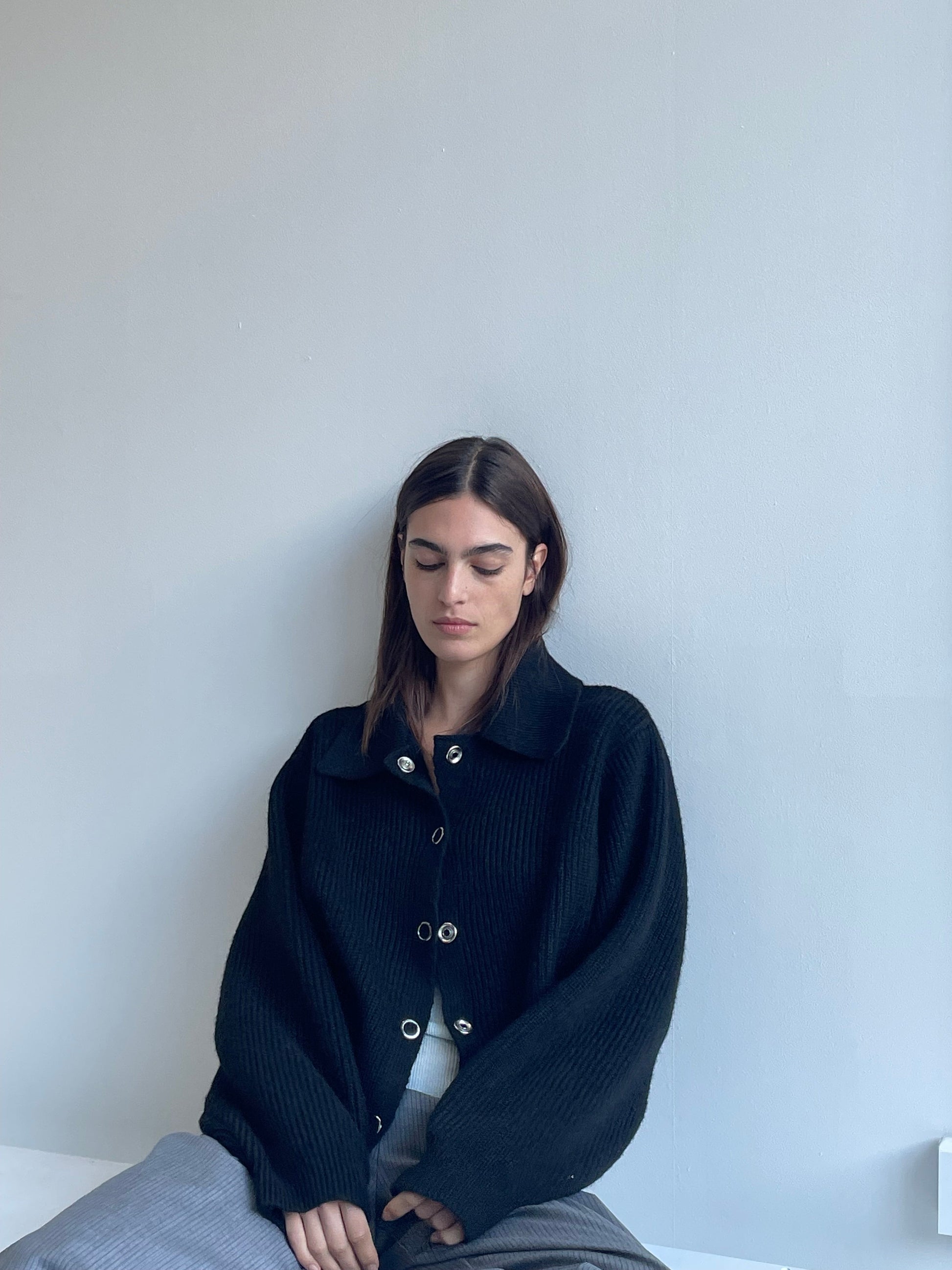 A woman with long brown hair sits against a light gray wall, wearing the Stud Cardi Knit - Black in an oversized fit and gray pants. She looks down, her expression calm and serene.