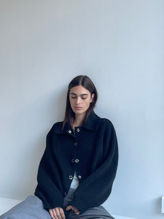A woman with long brown hair sits against a light gray wall, wearing the Stud Cardi Knit - Black in an oversized fit and gray pants. She looks down, her expression calm and serene.