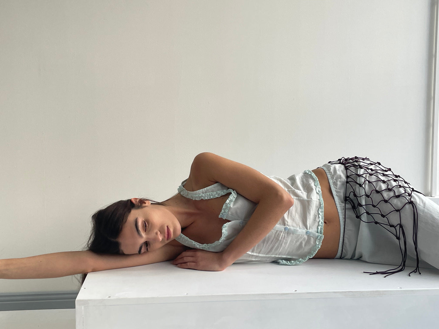 A woman in the Button Frill Top - Mint by Deiji Studios, made from organic cotton, lies on her side on a white surface, resting her head on her arm and appearing serene against a plain off-white background.