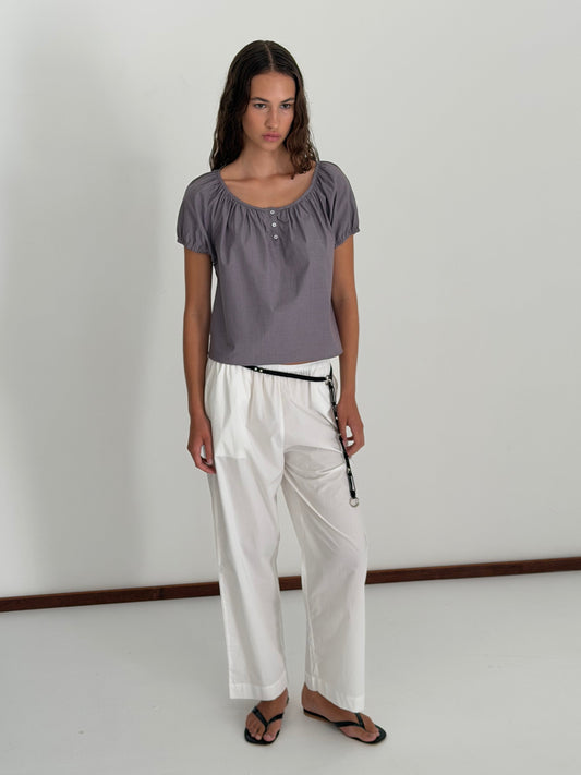 A woman with long wavy hair stands indoors against a white wall, wearing the Poplin T Shirt - Currant by Deiji Studios with loose white drawstring pants and black sandals, her expression neutral.