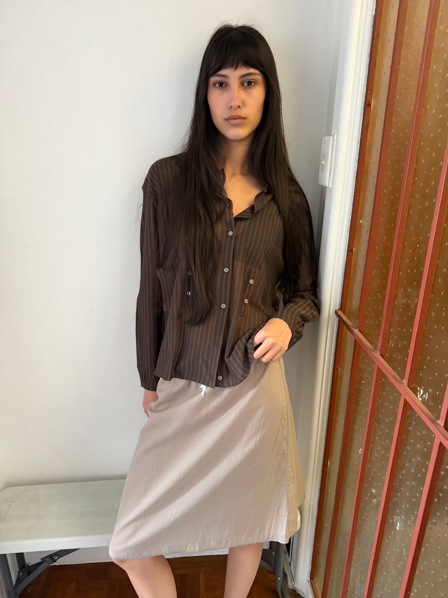A woman with long dark hair stands in the corner of a room wearing the Knit Shirt - Brown Grey Stripe by Deiji Studios, a sheer brown and charcoal striped knit shirt, over a pale skirt.