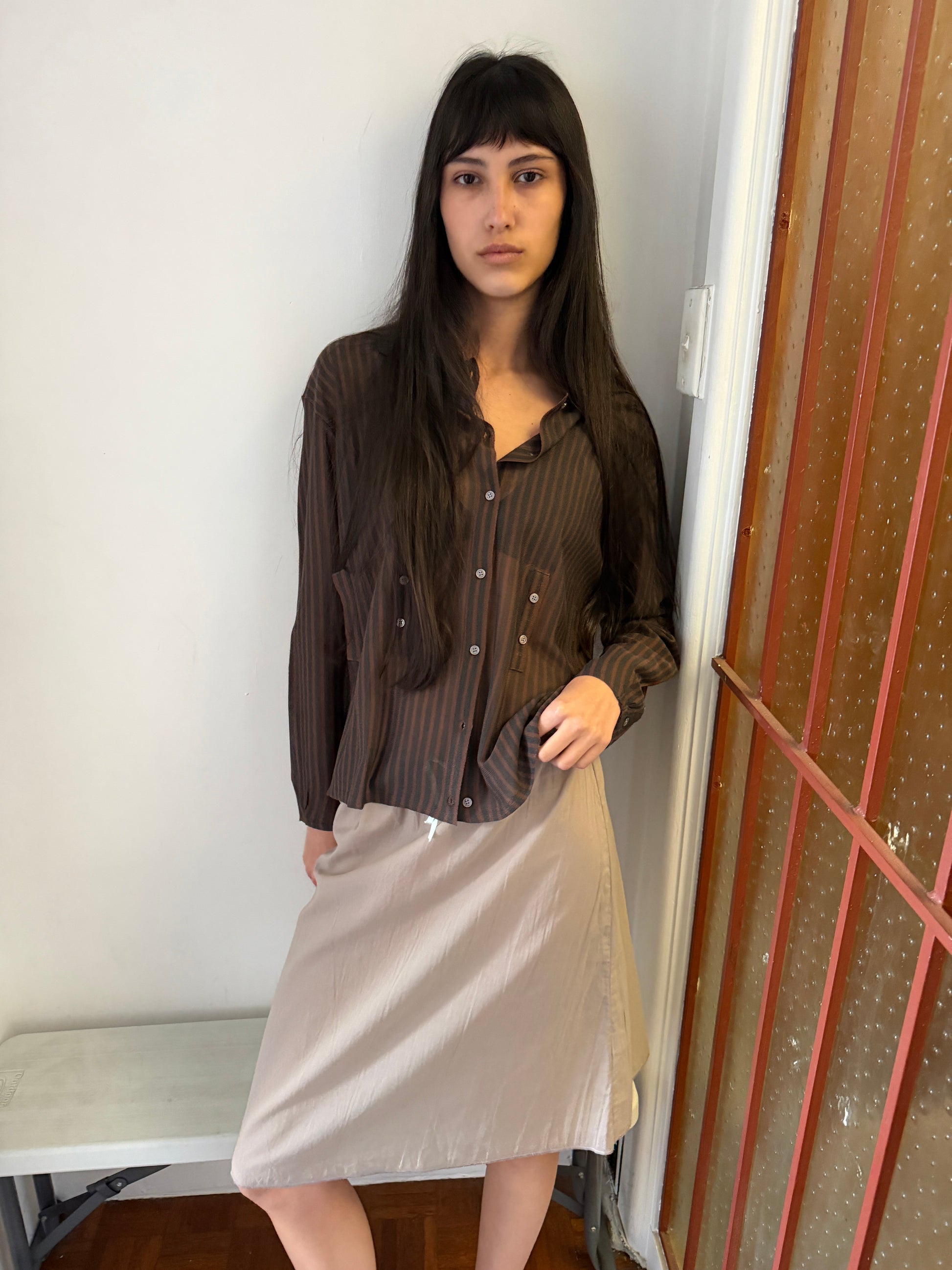 A woman with long dark hair stands in the corner of a room wearing the Knit Shirt - Brown Grey Stripe by Deiji Studios, a sheer brown and charcoal striped knit shirt, over a pale skirt.