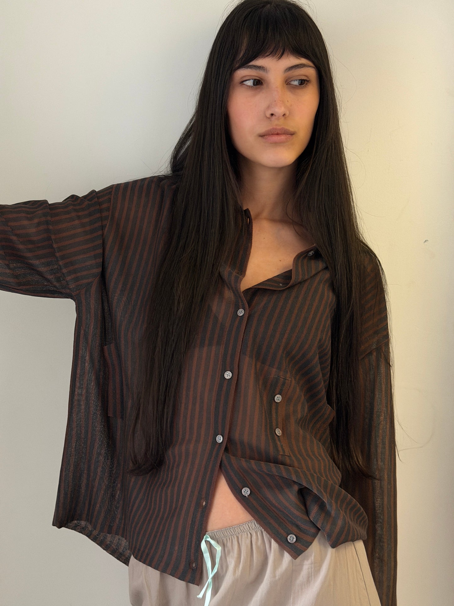 A woman with dark hair and a fringe leans against a wall wearing the Knit Shirt - Brown Grey Stripe by Deiji Studios, a sheer brown and charcoal striped knit shirt, over a cream drawstring skirt.