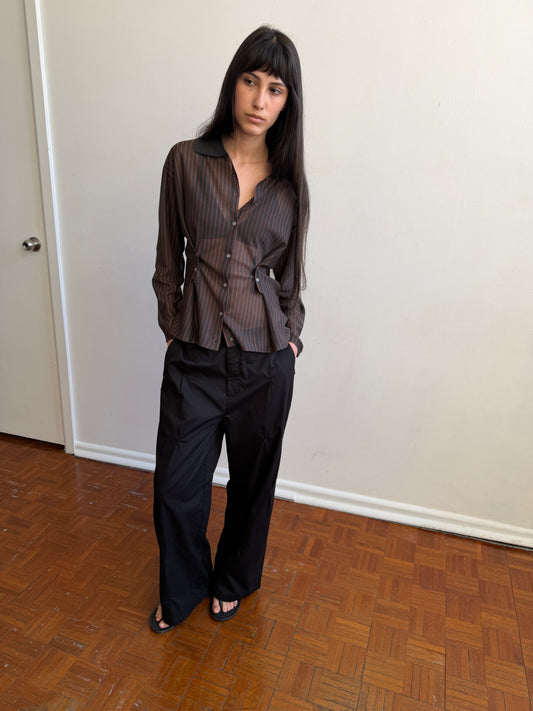 A woman with dark hair and a fringe stands in a room wearing the Multi Pleat Pant - Black by Deiji Studios, black wide-leg pleated trousers, with a sheer brown striped knit shirt and black sandals.