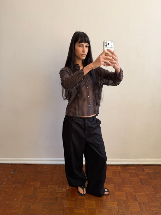 A woman with dark hair and a fringe takes a mirror photo wearing the Multi Pleat Pant - Black by Deiji Studios, black wide-leg pleated trousers, with a sheer brown striped knit shirt.