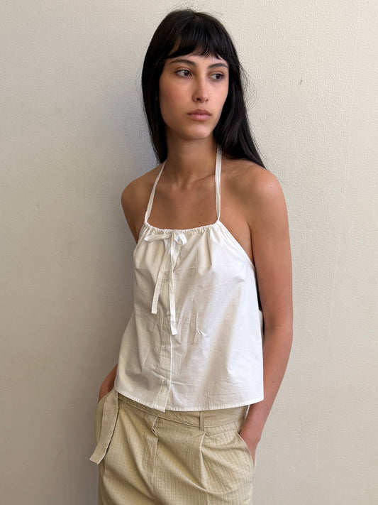 A cropped view of a woman with dark hair and a fringe wearing the Simple Tie Top - White by Deiji Studios, a white halter top with a gathered neckline and drawstring front tie, with cream trousers.