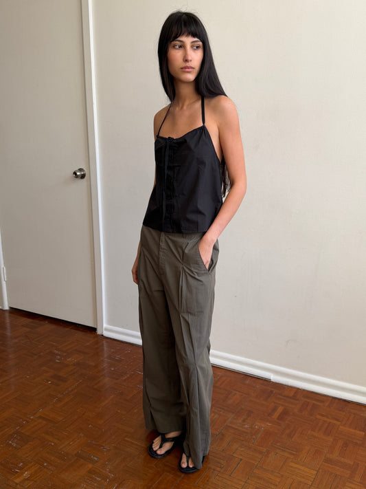 A woman with dark hair and a fringe stands in a room with a hand in her pocket, wearing the Multi Pleat Pant - Moss Stripe by Deiji Studios, moss-green fine-check wide-leg pleated trousers, with a black halter top and black sandals.