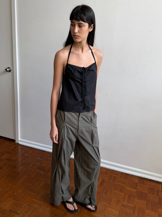 A woman with dark hair and a fringe stands in a room wearing the Multi Pleat Pant - Moss Stripe by Deiji Studios, moss-green fine-check wide-leg pleated trousers, with a black halter top and black sandals.