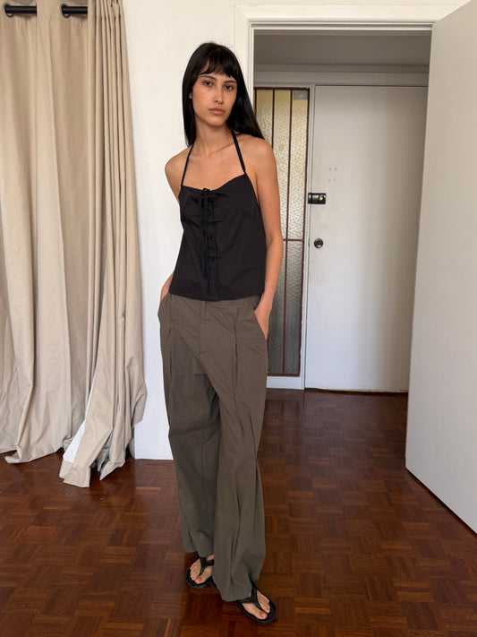 A woman with dark hair and a fringe stands in a room wearing the Simple Tie Top - Black by Deiji Studios, a black halter top with a gathered neckline and front tie, with khaki trousers and black sandals.