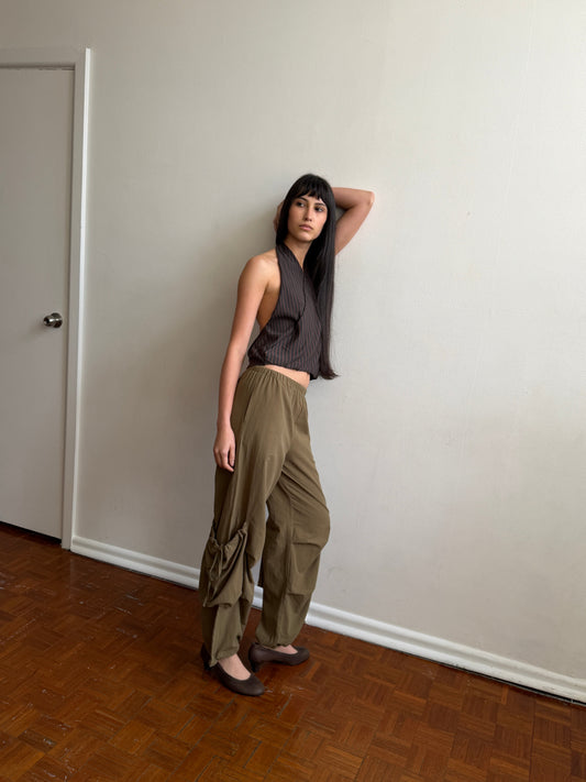 A woman with dark hair and a fringe leans against a wall wearing the Wrap Halter Top - Brown Grey Stripe by Deiji Studios, a sheer brown and grey striped halter top, with khaki drawcord trousers and brown flats.