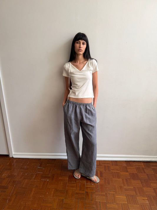 A woman with dark hair and a fringe stands against a wall wearing the Linen Ease Trouser - Indigo Check by Deiji Studios, indigo micro-check wide-leg linen trousers, with a cream tie-back tee and grey flip-flops.