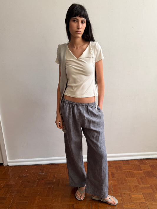 A woman with dark hair and a fringe stands in a room wearing the Linen Ease Trouser - Indigo Check by Deiji Studios, indigo micro-check wide-leg linen trousers, with a cream tie-back tee and grey flip-flops.